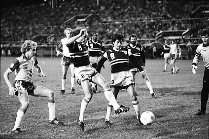 Black-and-white photo of a men’s soccer team kicking a ball toward the goal of their opponents, as the defending team tries to clear the ball away from the front of the net. Spectators form a blur in the stands in the background.