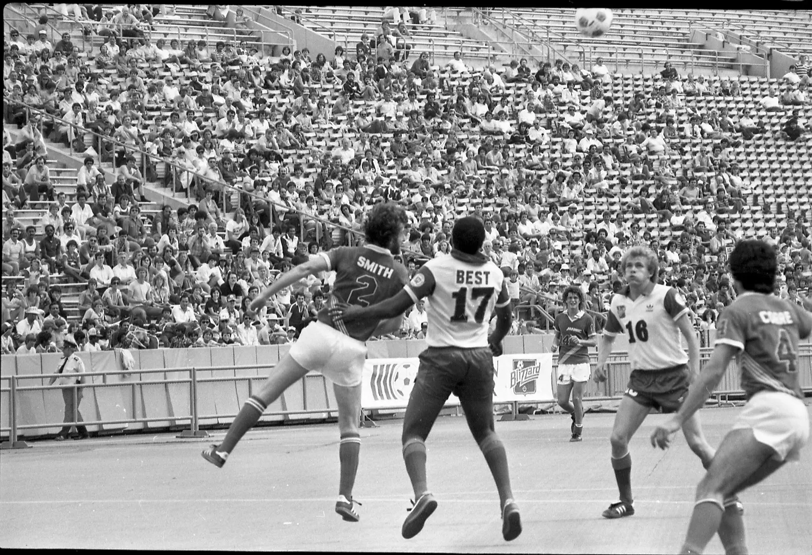 Black-and-white photo of two soccer players vying to get a better position on an incoming ball. There are spectators seated and standing in bleachers in the background.