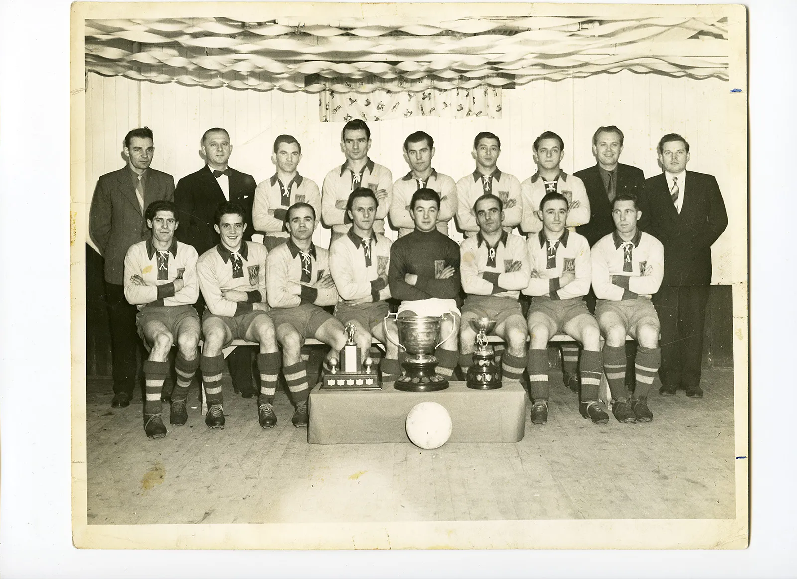 Yellowed black-and-white photo of a soccer team arranged in two rows, shot indoors. The team lines up behind the three trophies they have just won, and there appear to be streamers hung on the ceiling.