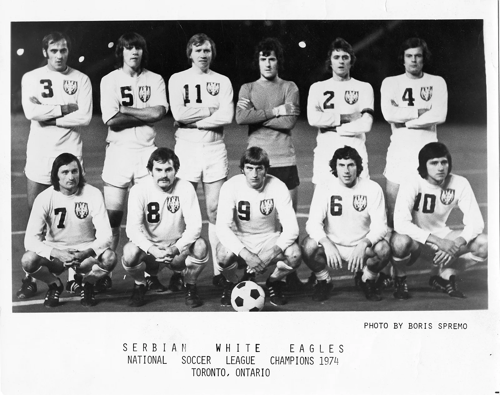 Black-and-white photo of a soccer team on a soccer field. Players wear white uniforms with insignia on them.