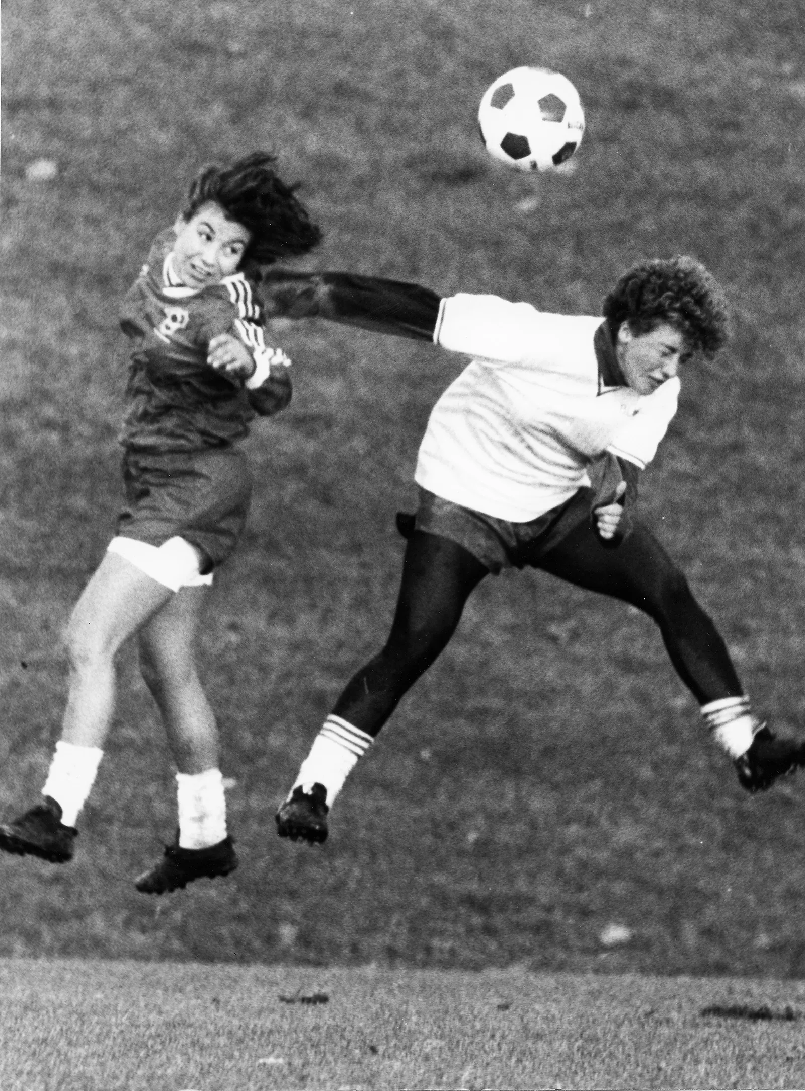 Black-and-white photo of a soccer player performing a header against a player from the opposing team. Both opponents appear to give their full efforts.