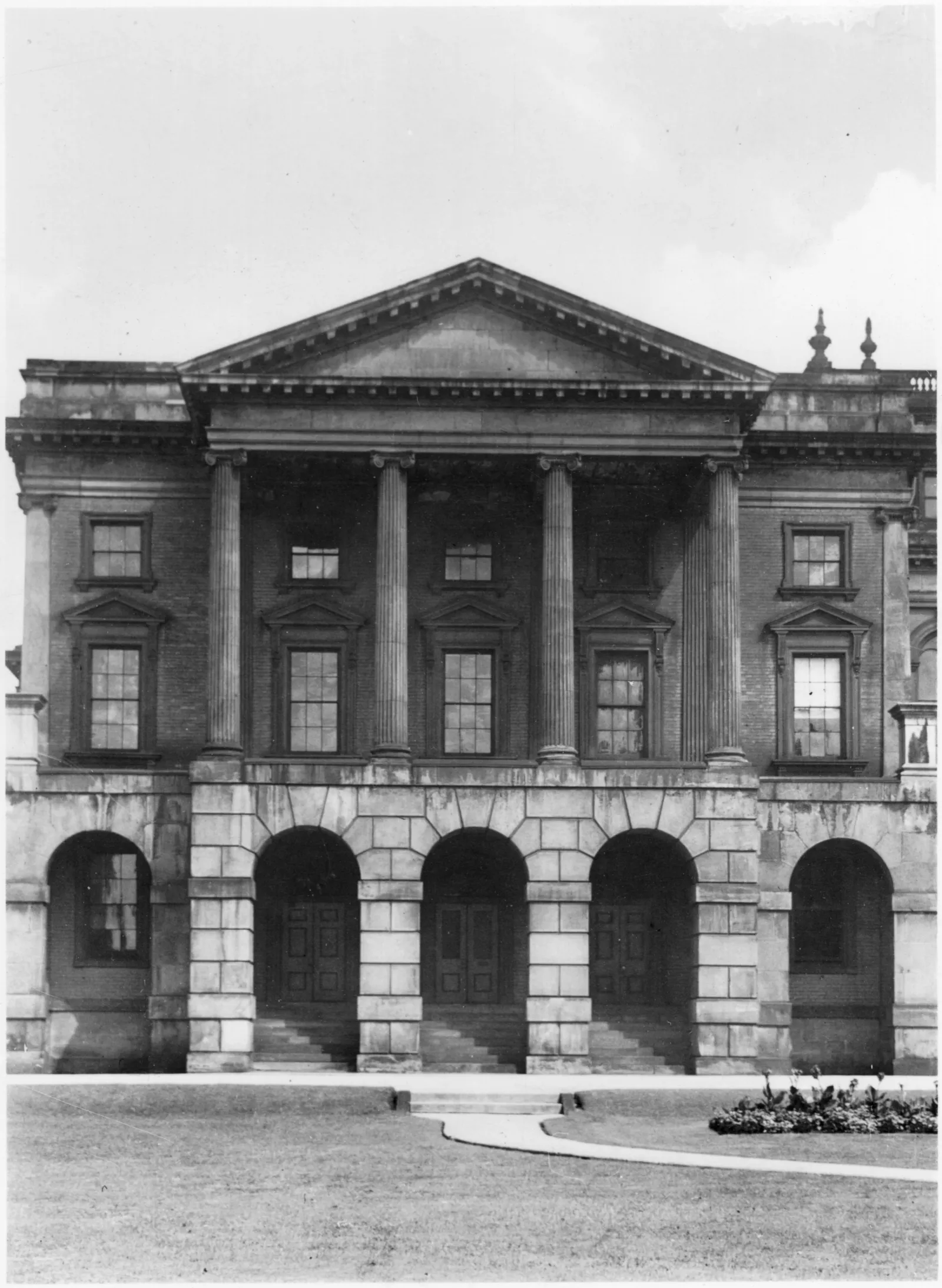Black-and-white photograph showing the front elevation of the West Wing. The building features a portico with a large triangular pediment and four tall columns, set atop a ground floor entrance with stone arches.