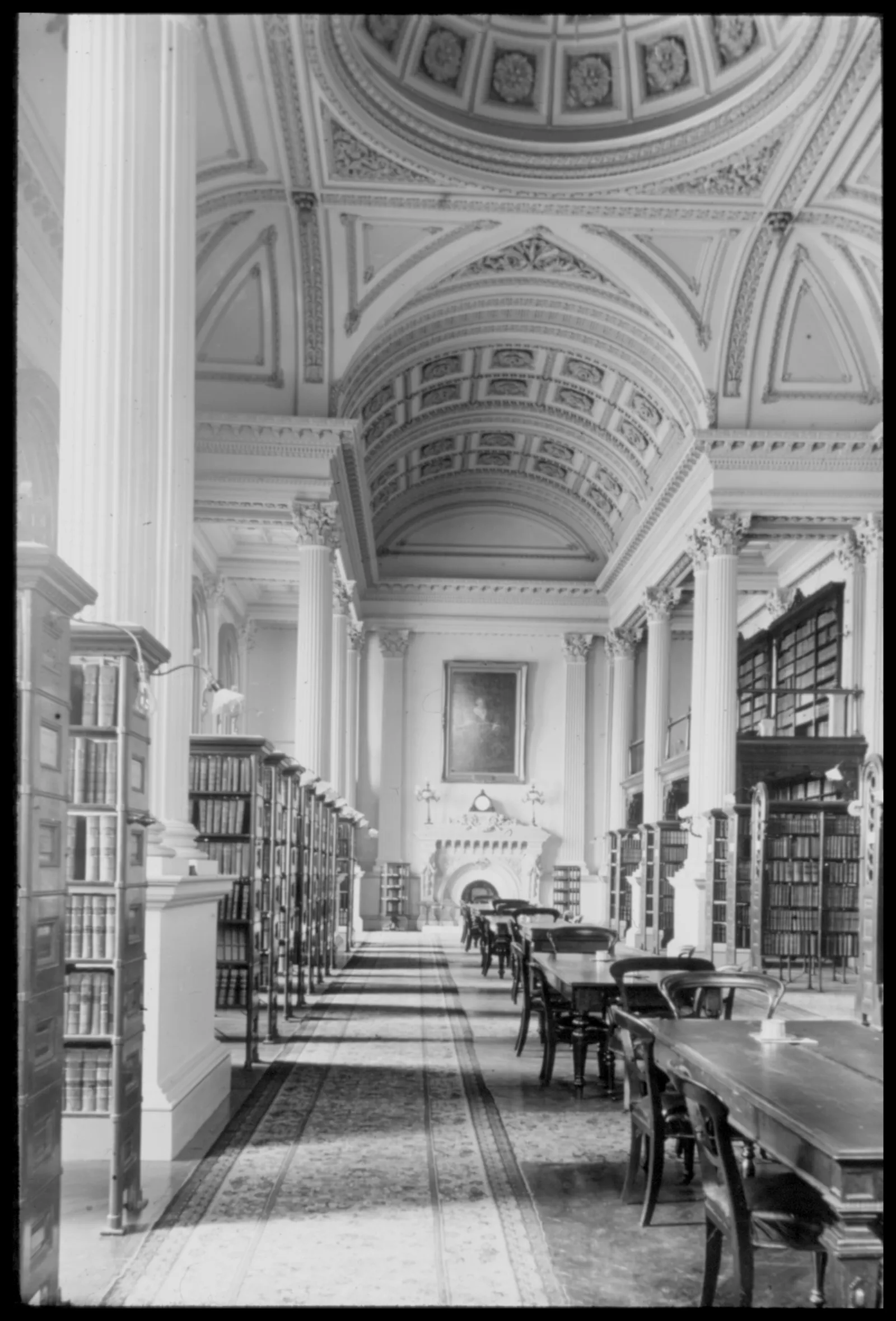 Black-and-white photograph of the interior of the Great Library featuring high vaulted ceilings and grand columns. Long rows of tall wooden bookshelves and reading tables are arranged throughout the hall, leading toward a fireplace and a portrait at the end of the hall.