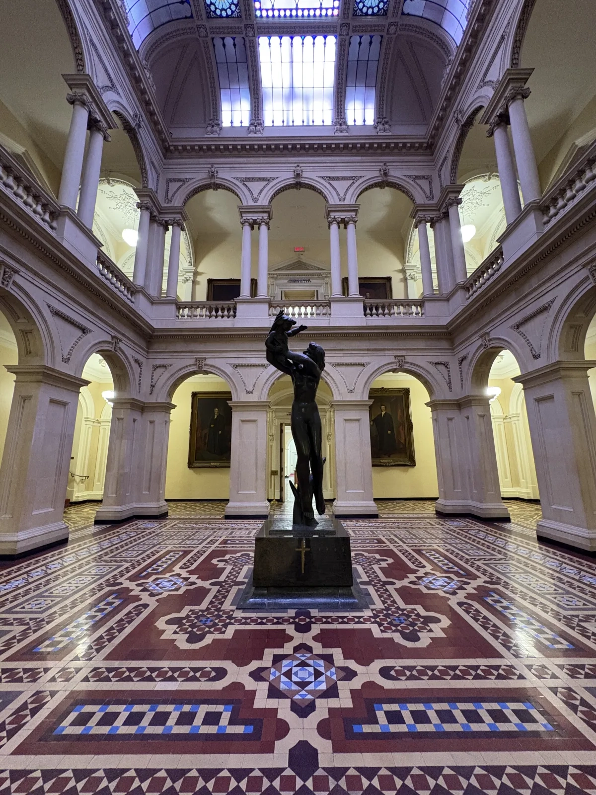 Photograph showing a large atrium with a bronze statue of a woman holding a child at the centre, set on an intricate geometric tiled floor. Arches and a large decorative skylight define the two-level space, while framed portraits line the surrounding gallery walls.