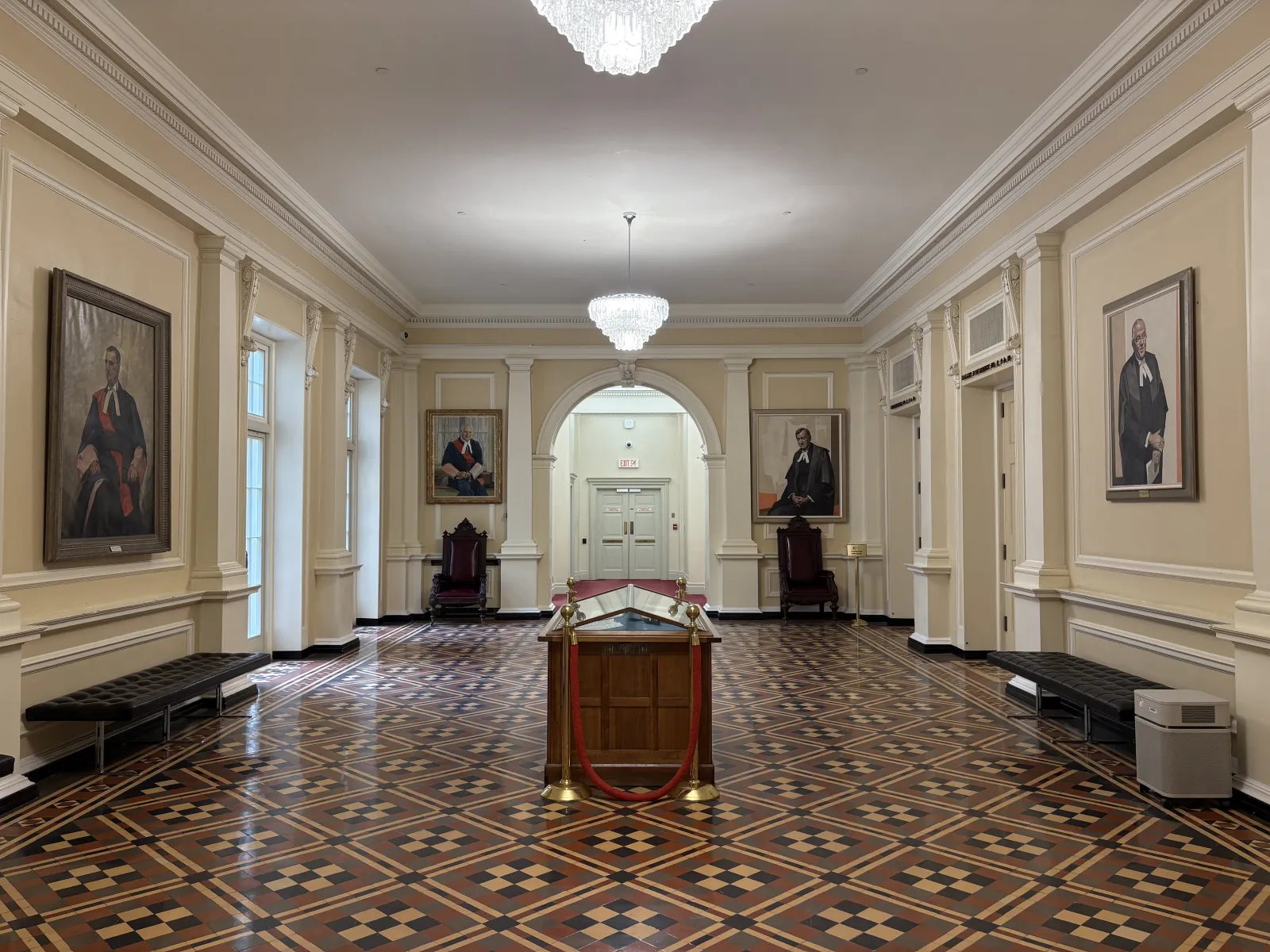 Photograph of a large room with a detailed geometric tiled floor and walls lined with framed portraits and columns. Crystal chandeliers illuminate the room, which contains black leather benches and a central wooden display case.