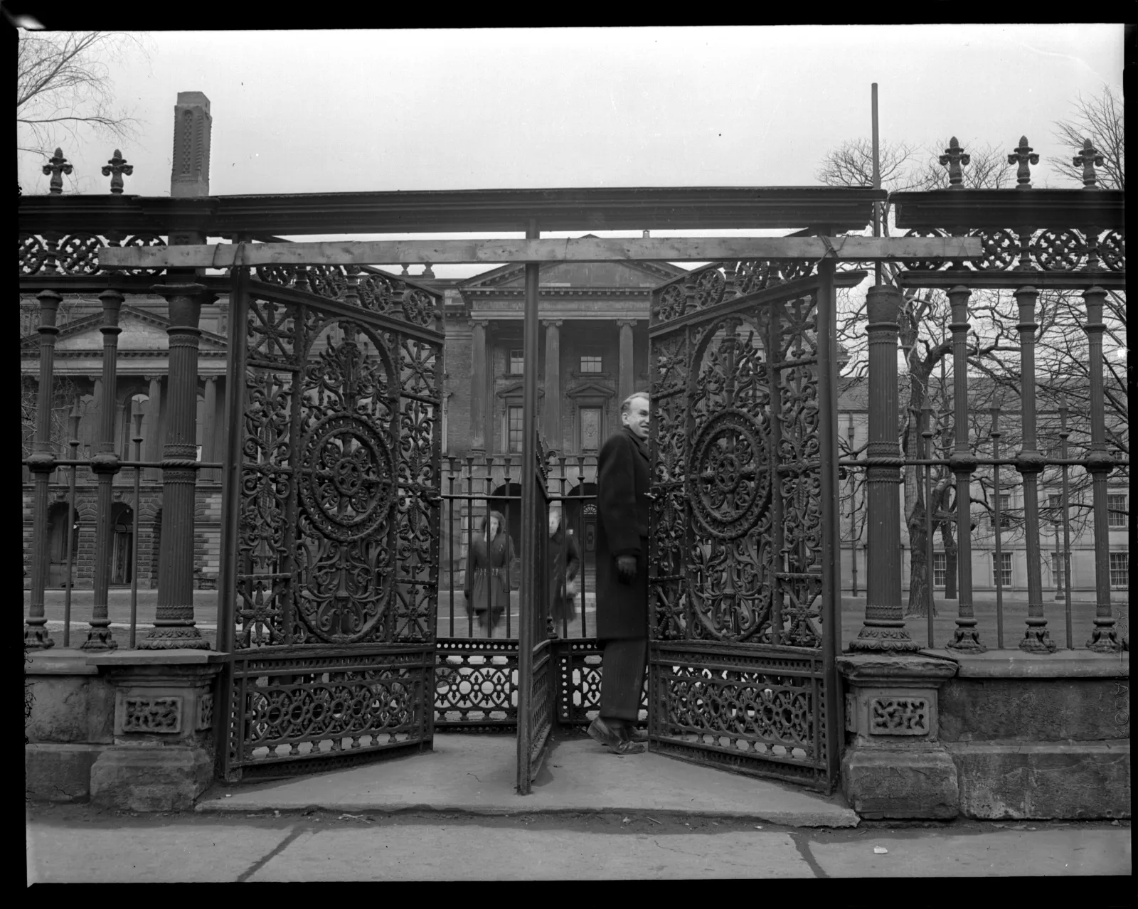 Black-and-white photograph depicting three people at the cast-iron gates of Osgoode Hall. A man in a long coat stands in the foreground, partially passing through the open squeeze gate, while two women are visible behind the gates in the mid-ground. In the background, the grand facade of Osgoode Hall is visible through the iron bars.
