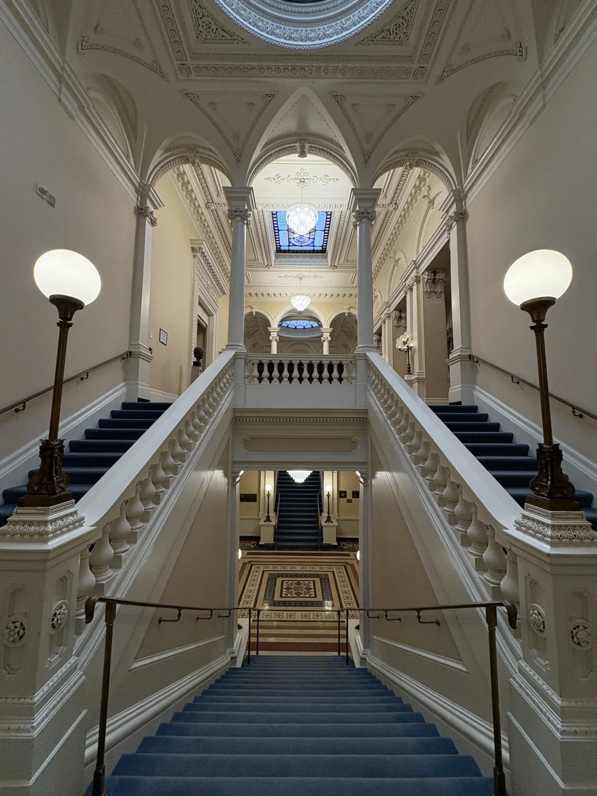 Photo of a symmetrical grand staircase featuring blue-carpeted steps, white arches and globe lamps. An intricate rectangular skylight illuminates the decorative ceiling mouldings of a space visible at the top of the staircase.