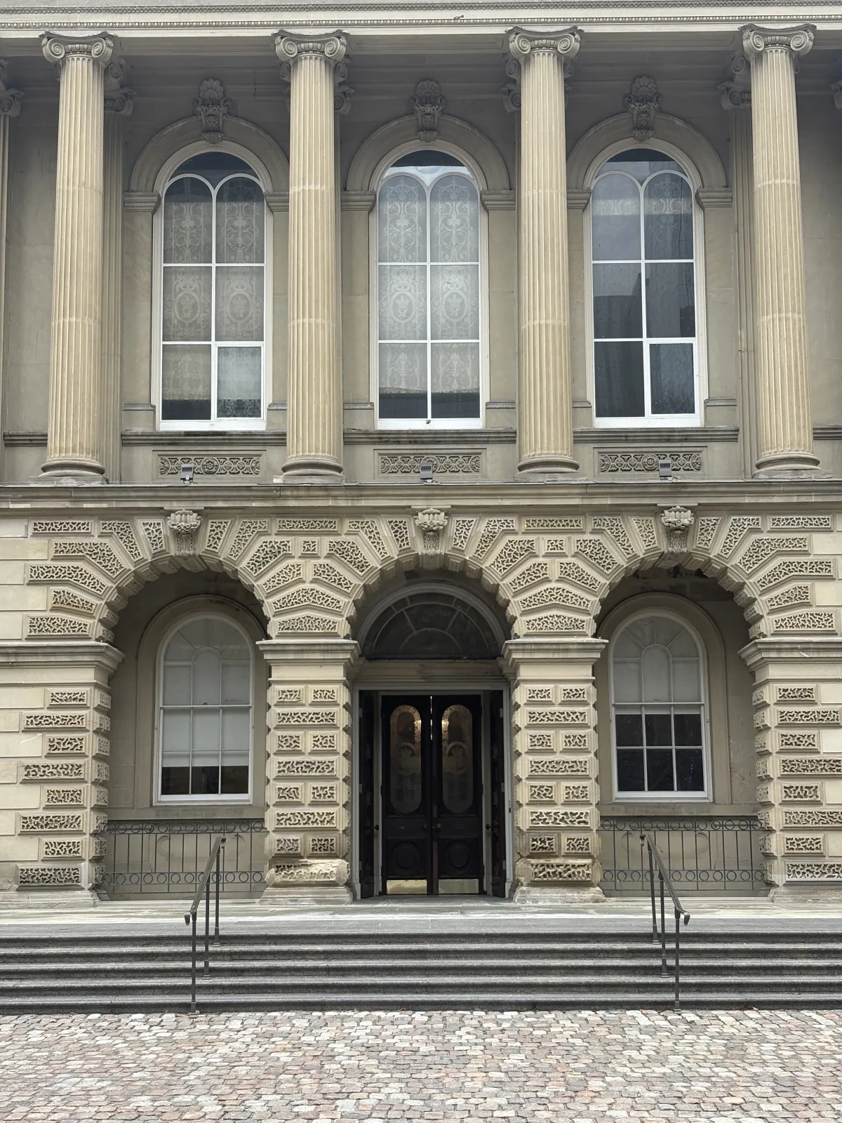 Photograph showing the centre entrance of Osgoode Hall, featuring three grand arches with intricate stone rustication and four fluted columns. A dark wooden doorway is centred at the top of the stone steps.