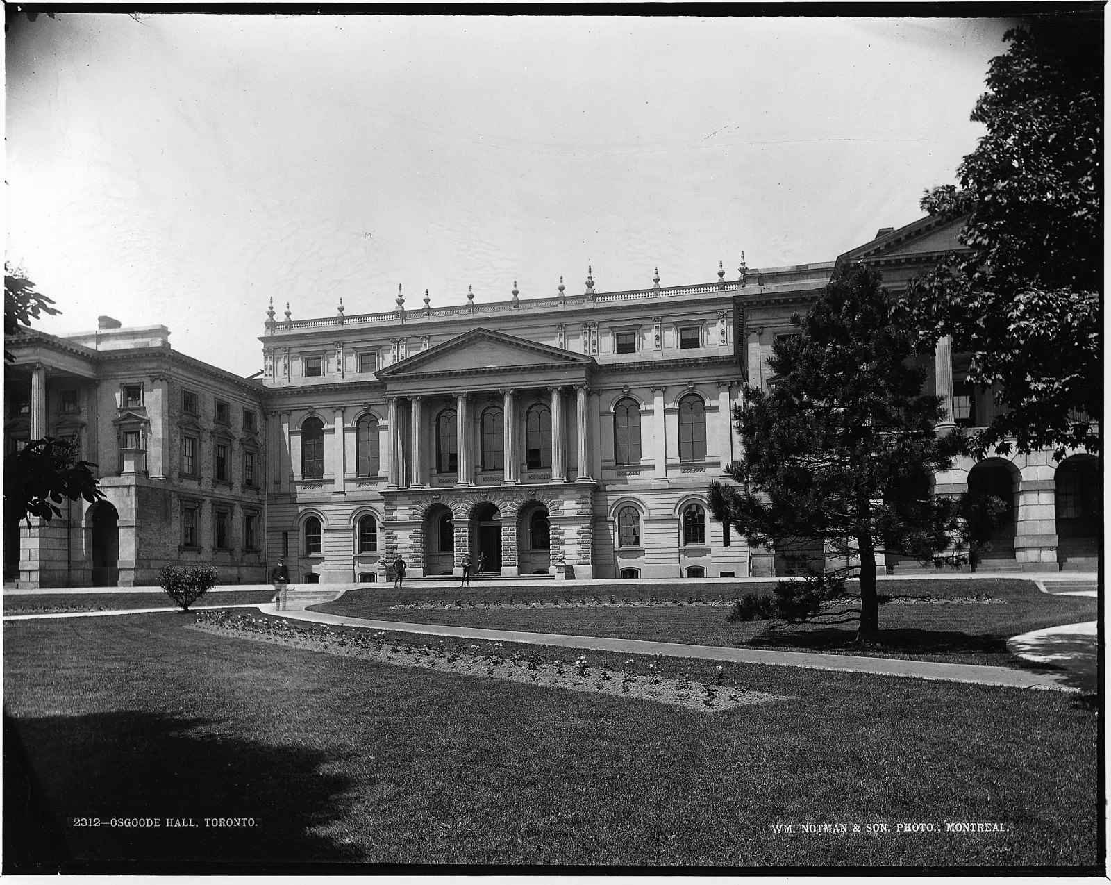 Black-and-white photograph showing the south facade of Osgoode Hall featuring a manicured lawn and decorative garden beds in the foreground. The west part of the building shows deterioration of the paint coating on the original brick walls.