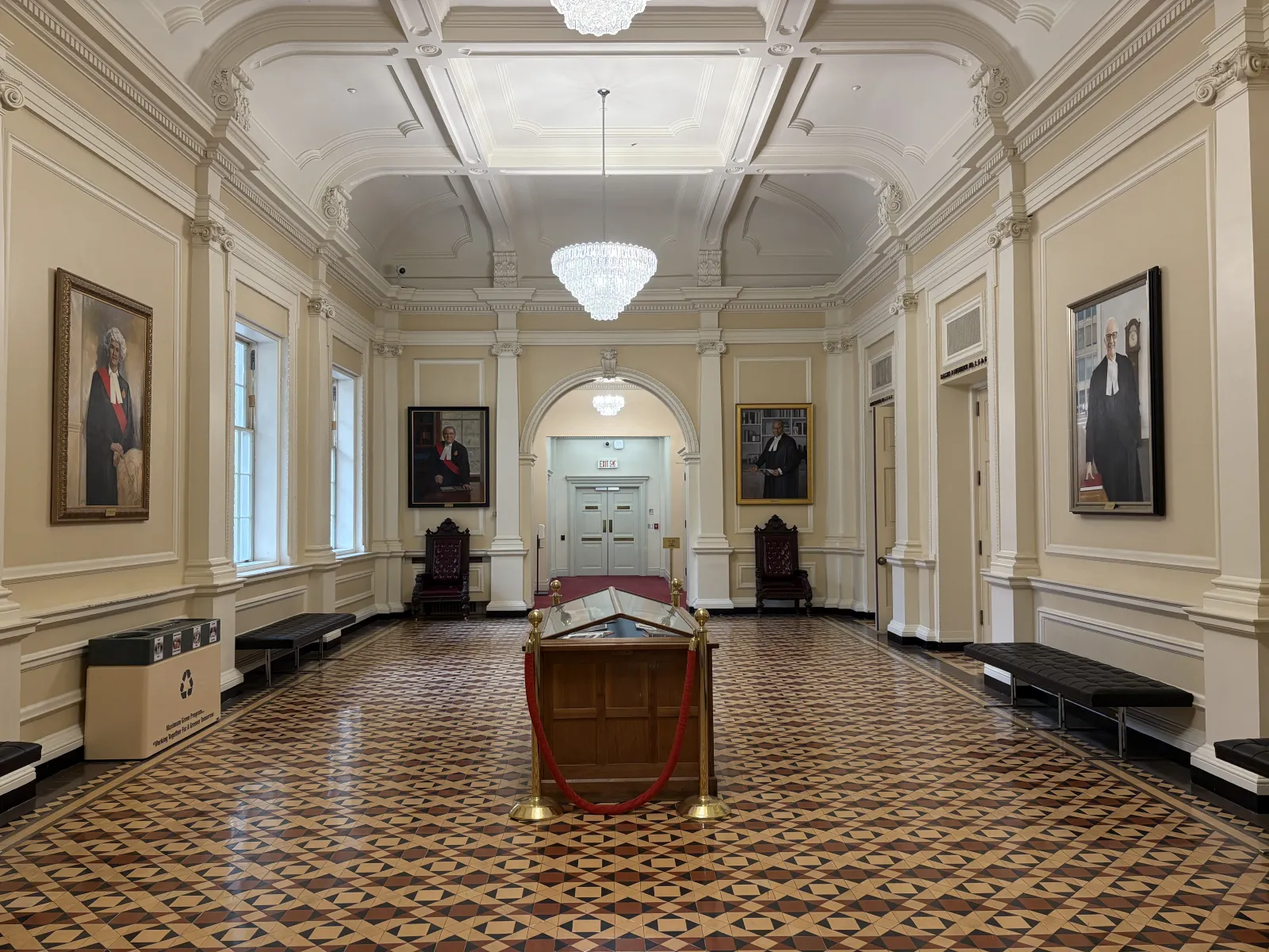 Photograph of a large room with a detailed geometric tiled floor and walls lined with framed portraits and columns. Crystal chandeliers illuminate the room’s ornate ceiling, and black leather benches and a central wooden display case are placed along the walls.