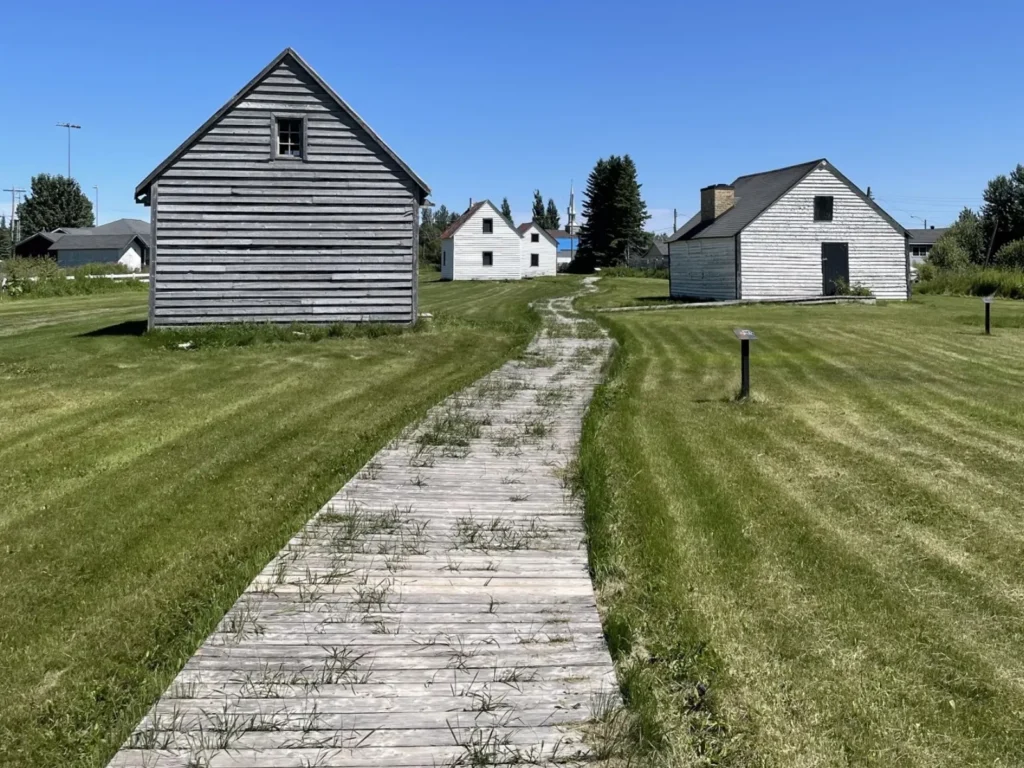 A wooden boardwalk leads through a grassy heritage site with several historic white clapboard buildings under a clear blue sky.