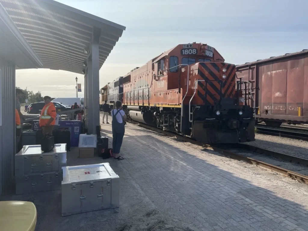 A red locomotive is stopped beside a train station platform, with large metal cases, rail staff, and visitors gathered nearby.