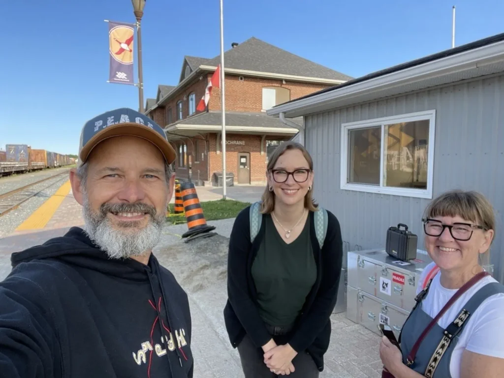Three people pose for a selfie on a railway platform in front of a brick train station building. Train tracks and a freight train run along the left side of the image, while equipment cases and a small gray building are visible on the right under a clear blue sky.