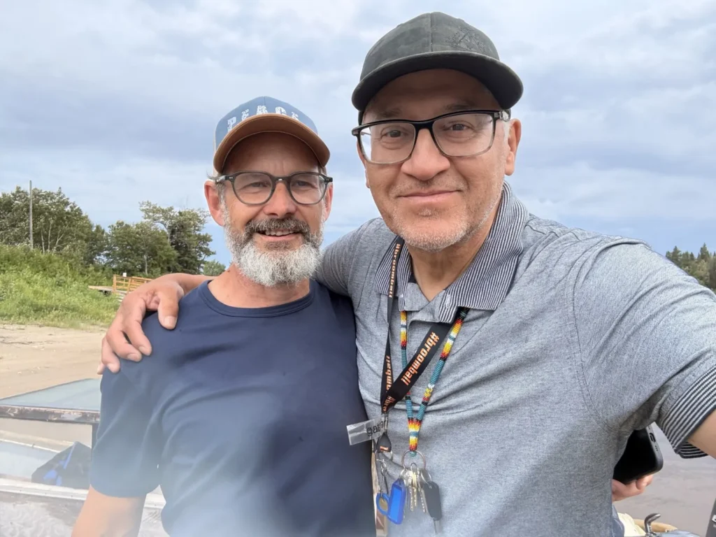 Two people wearing glasses and baseball caps stand outdoors with their arms around each other, posing for a photo under a cloudy sky.