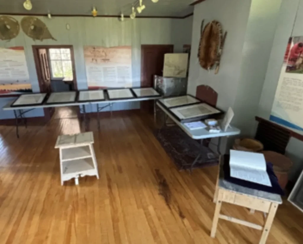 Historic museum room with polished wooden floors, display tables containing documents and artifacts, informational wall panels, and wooden stools arranged throughout the exhibit space.