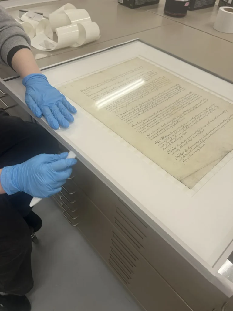 A historical handwritten document being prepared for preservation and display in a glass-covered frame, with gloved hands working to adjust the frame in a conservation workspace.