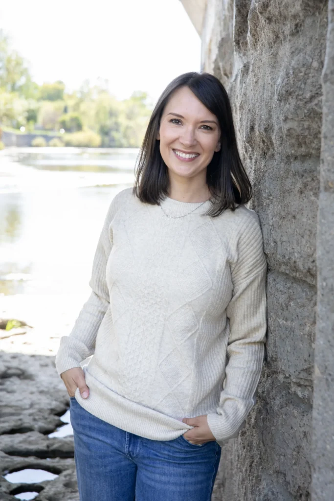 Outdoor portrait of a woman standing beside a stone wall near a river or waterfront. She wears a light-coloured knit sweater and blue jeans, with her hands in her pockets. Trees, water, and a bridge structure are visible in the softly blurred background.