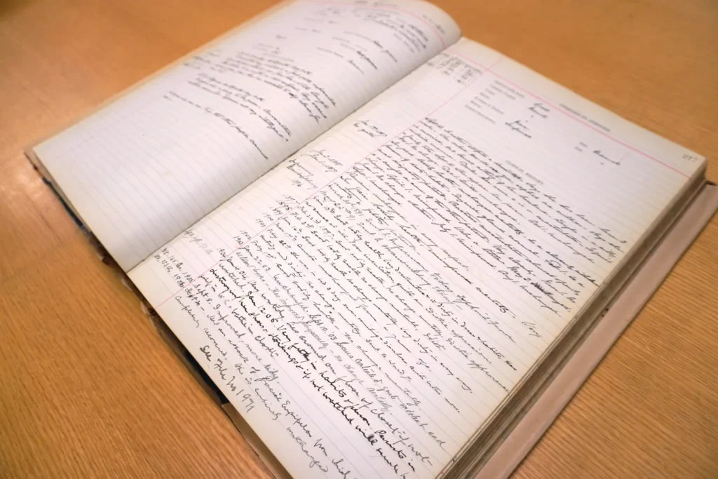 Open archival ledger book resting on a wooden table. The pages contain handwritten entries in ink arranged in ruled columns, with dates, names and notes recorded across both pages. The bound volume is opened near the middle, revealing detailed historical record-keeping and administrative documentation.