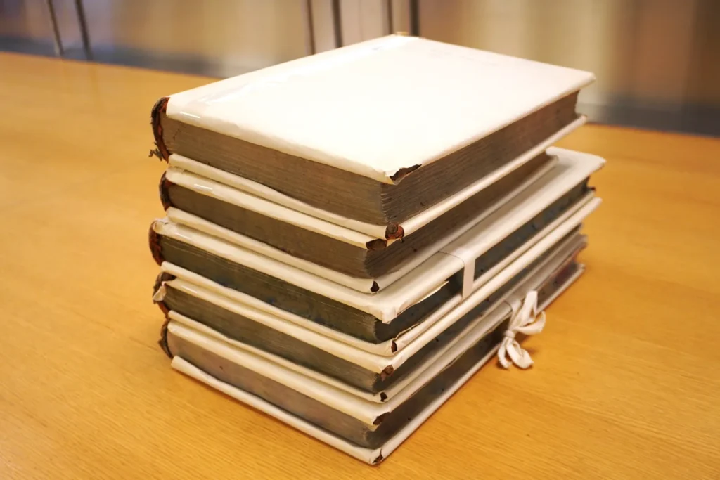 Stack of five large bound archival volumes tied with white protective paper wrapping and twill tape, resting on a wooden table. The worn book edges and thick pages indicate frequent use and long-term preservation of historical records within an archival collection.