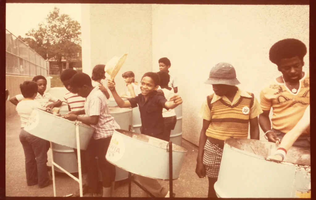 Colour photograph of a steelpan band performing on a float or raised platform during a street festival or parade. Several musicians in matching yellow shirts play light blue steel drums mounted on stands, while spectators gather below. Urban buildings and a large billboard are visible in the background, capturing a lively outdoor performance in a city setting.