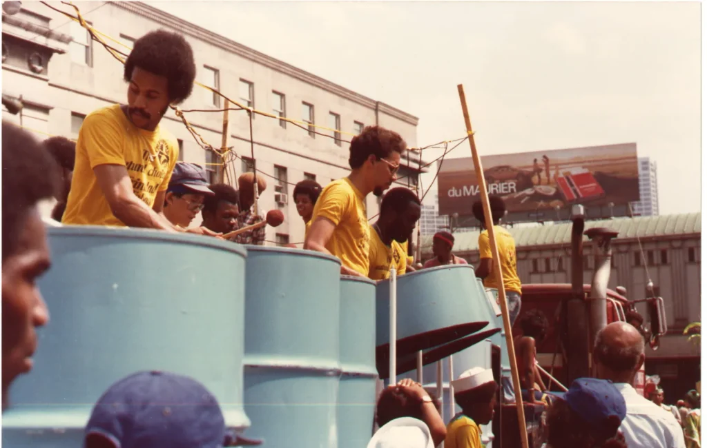 Colour photograph of a steelpan band performing on a float or raised platform during a street festival or parade. Several musicians in matching yellow shirts play light blue steel drums mounted on stands, while spectators gather below. Urban buildings and a large billboard are visible in the background, capturing a lively outdoor performance in a city setting.