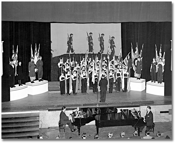 Photographie : Eaton’s employees’ War Bond Rally; women on stage dressed as ordnance workers, holding flags, singing, 1943