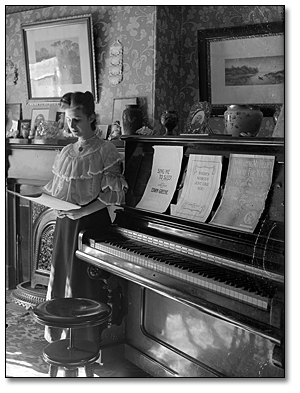 Unidentified woman looking at sheet music, posed beside a piano, [between 1898 and 1920]