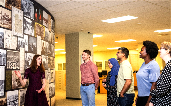 Woman 
gesturing towards a wall of images depicting archival records in front of a group of five smiling tour participants.