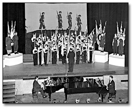 Photographie :  Eaton’s employees’ War Bond Rally; women on stage dressed as ordnance workers, holding flags, singing, 1943
