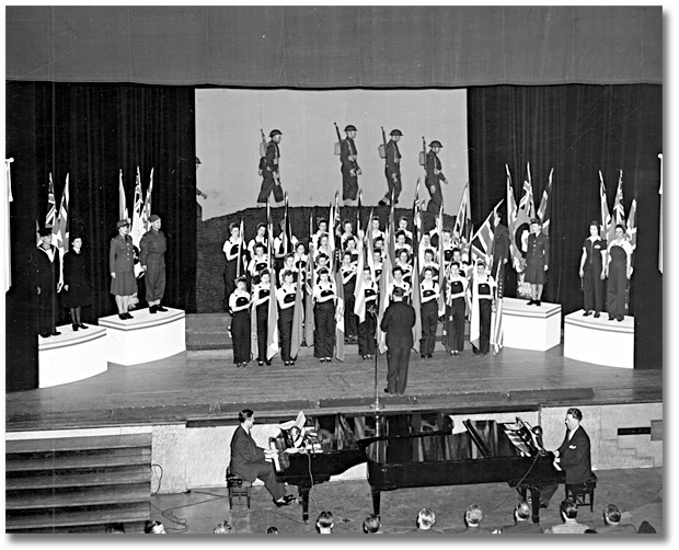 Photo: Eaton’s employees’ War Bond Rally; women on stage dressed as ordnance workers, holding flags, singing, 1943