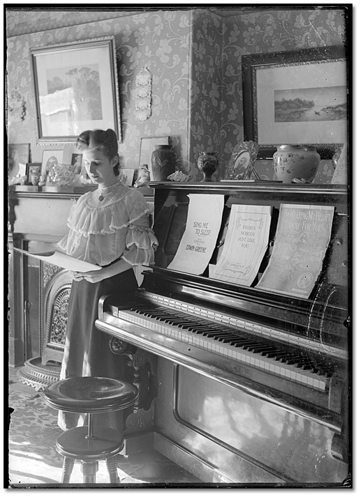 Photo: Woman looking at sheet music, posed beside a piano, [between 1898 and 1920]