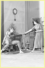 Click for Two white actors on stage. A woman pulls on a rope around a man tied to a chair.